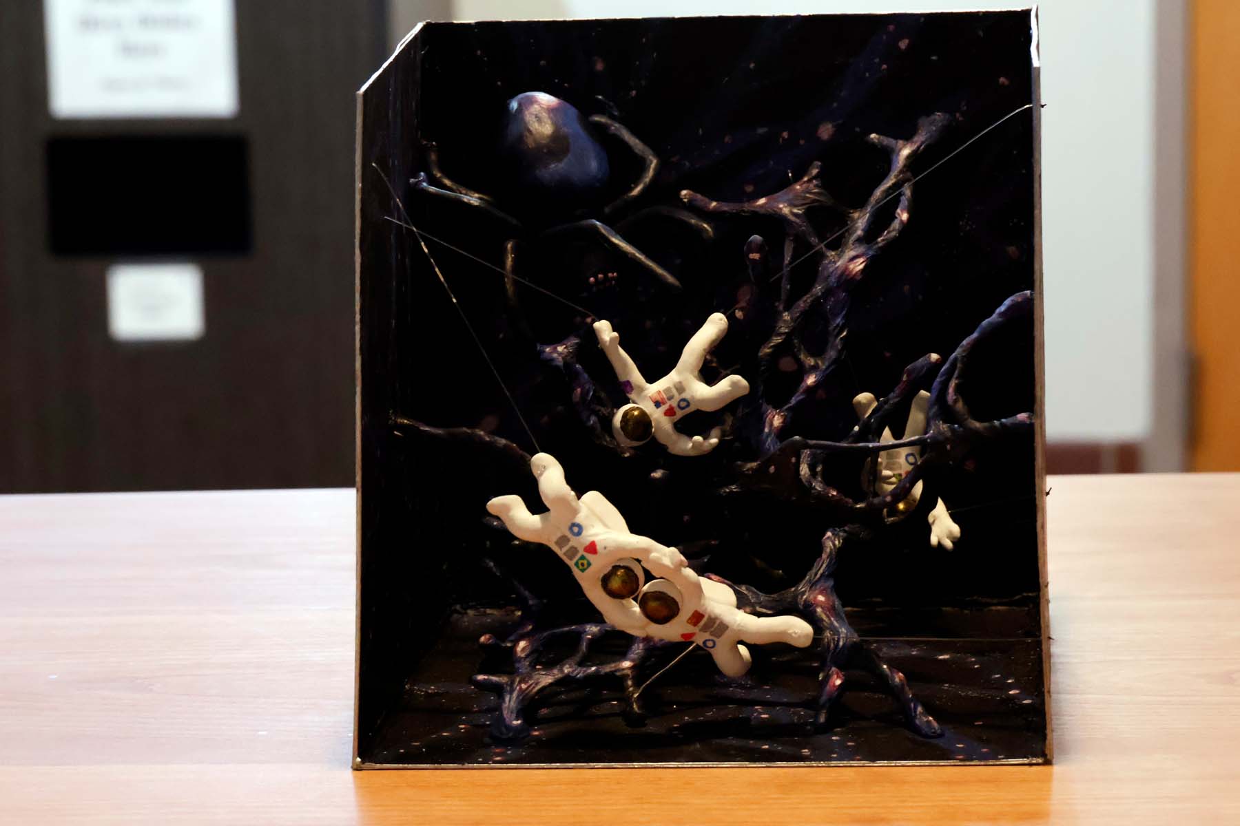 A cube, which is open on two sides (one opening facing us, the other facing above), sits on a table. The inside walls are painted with stars and between them stretch long, web-like filaments that carry the same starry pattern. In the upper left of the box, a cosmically colored spider straddles these threads while four astronauts float near various portions of the "web". The astronauts in the foreground are clutching each other's elbows, with helmets resting together; the astronauts are colinear such that a line would pass through the first's feet, then their touching helmets, then exit at the second's feet.