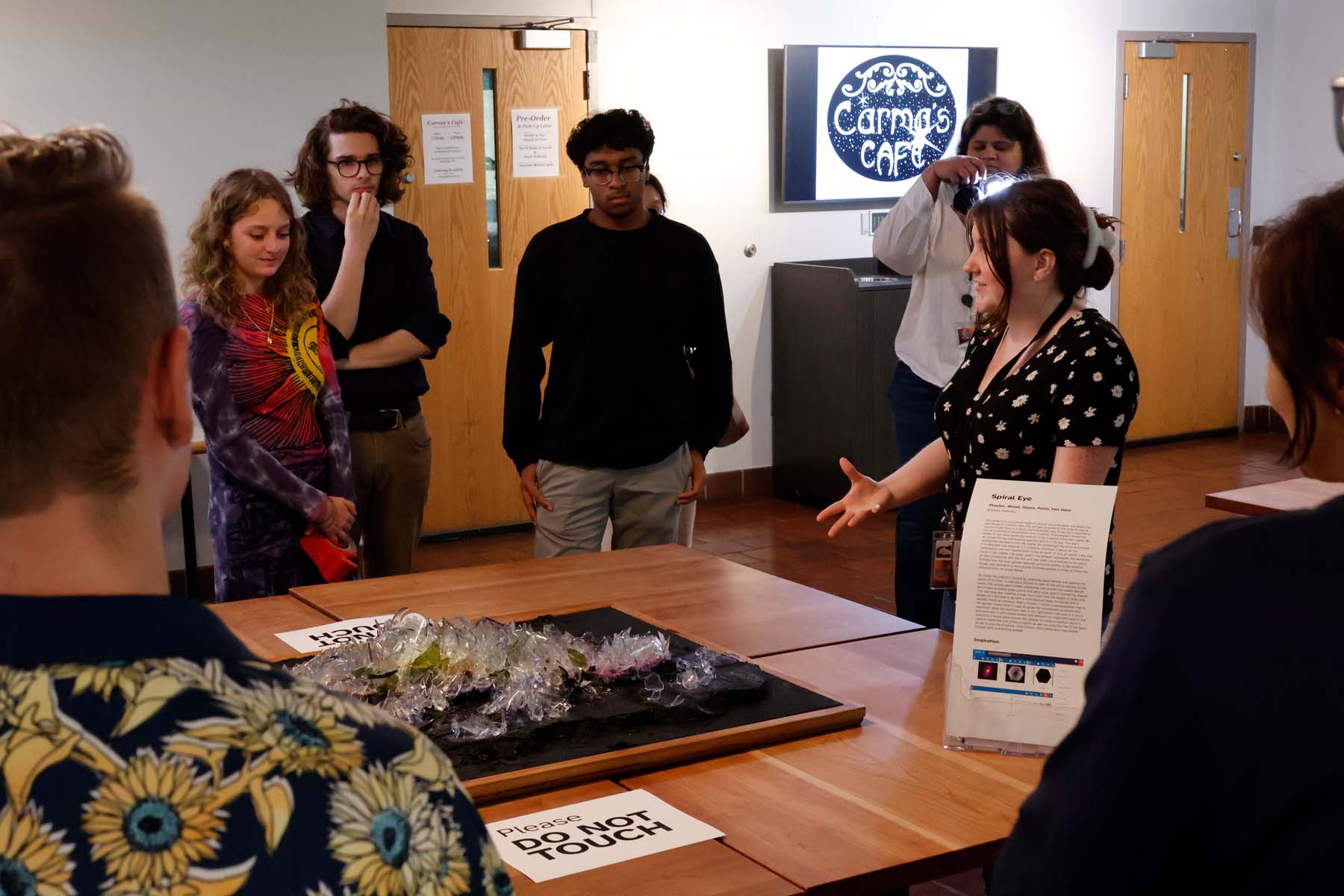 A student motions with her hand as she describes her sculpture to a group of onlookers. The sculpture sits on a table and is made from broken glass pieces formed into the shape of a spiral galaxy.