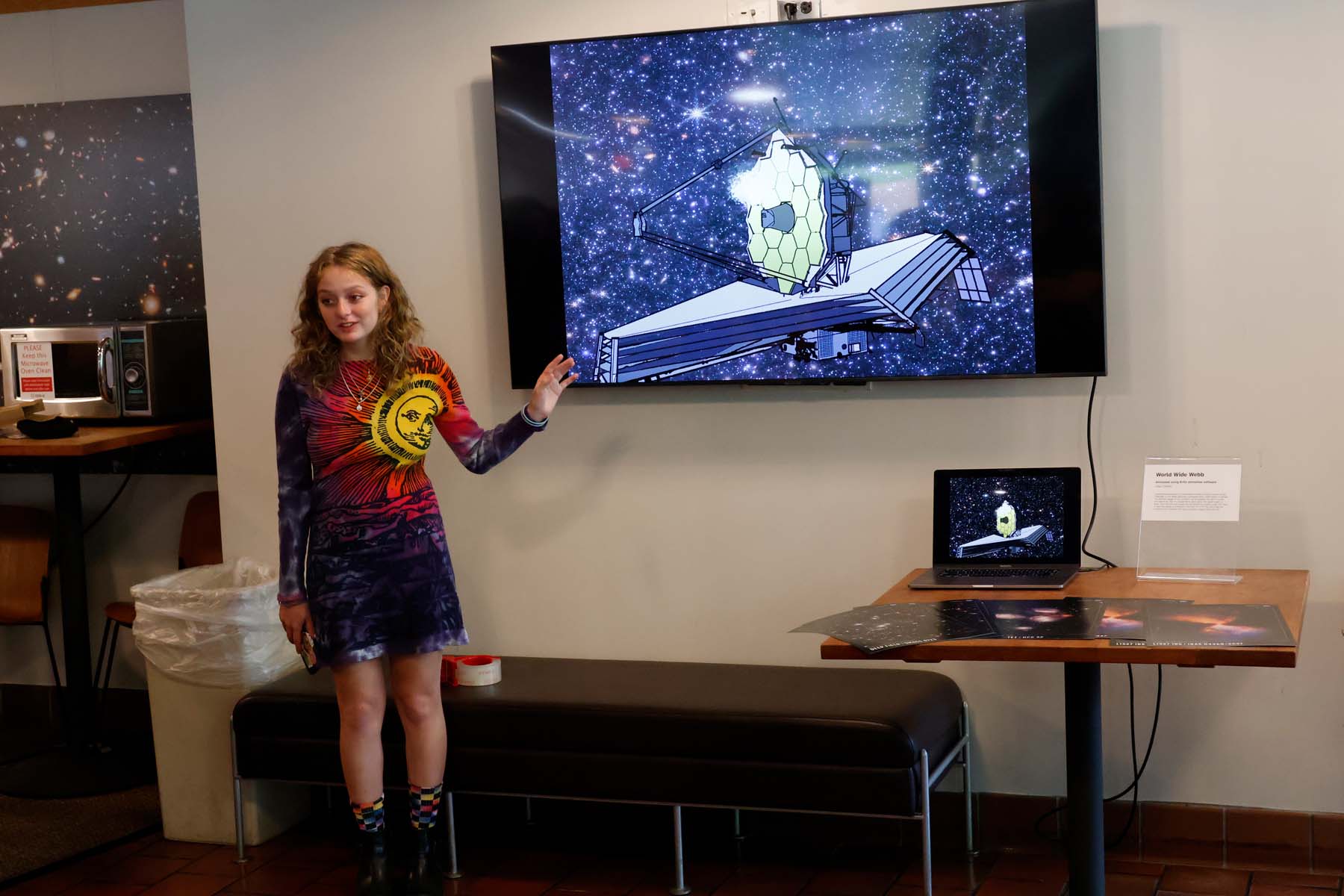 A student wearing a dress with the sun printed on it motions towards a large television screen playing her animation of the James Webb Space Telescope.