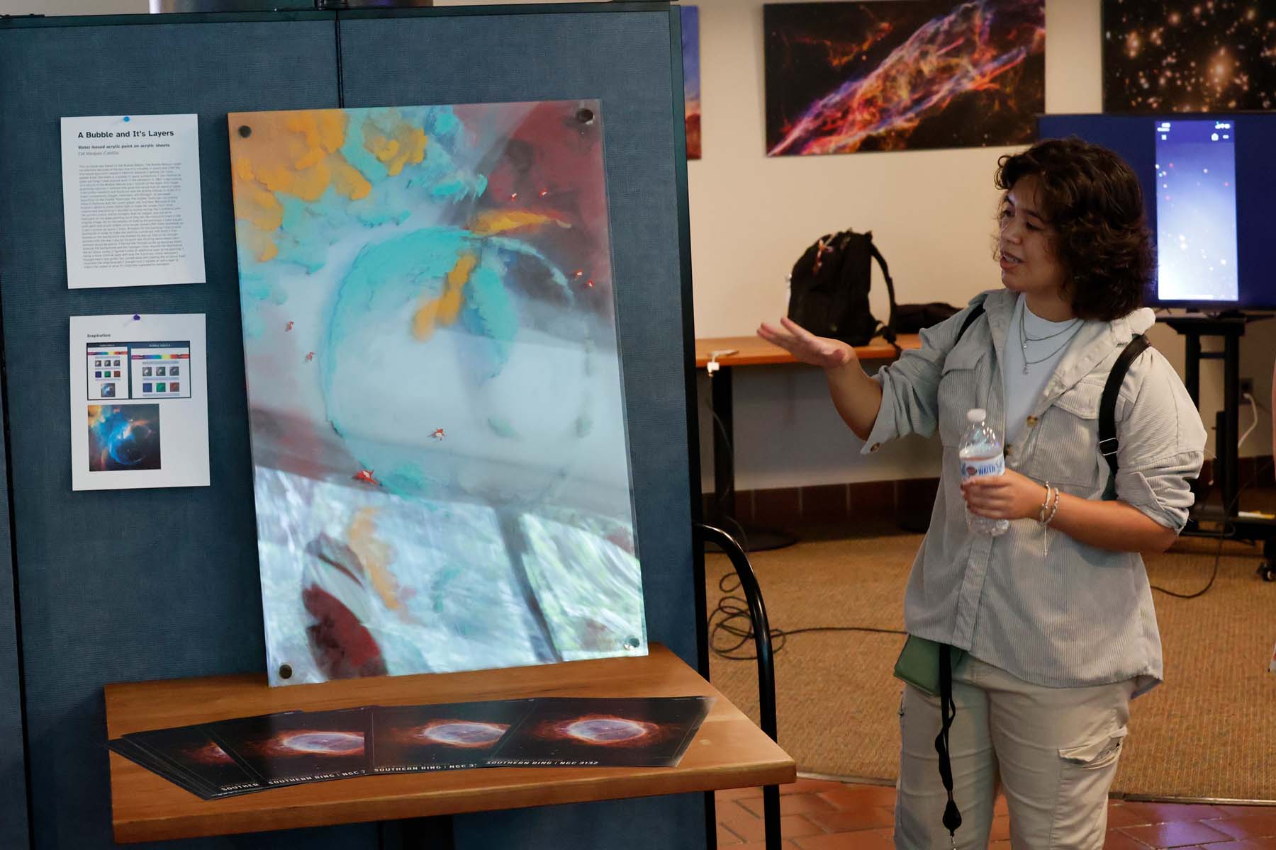 A student, caught mid-explanation, shows off a painted bubble nebula; the piece is layered plexiglass that has been painted and stacked to give it a textured look.