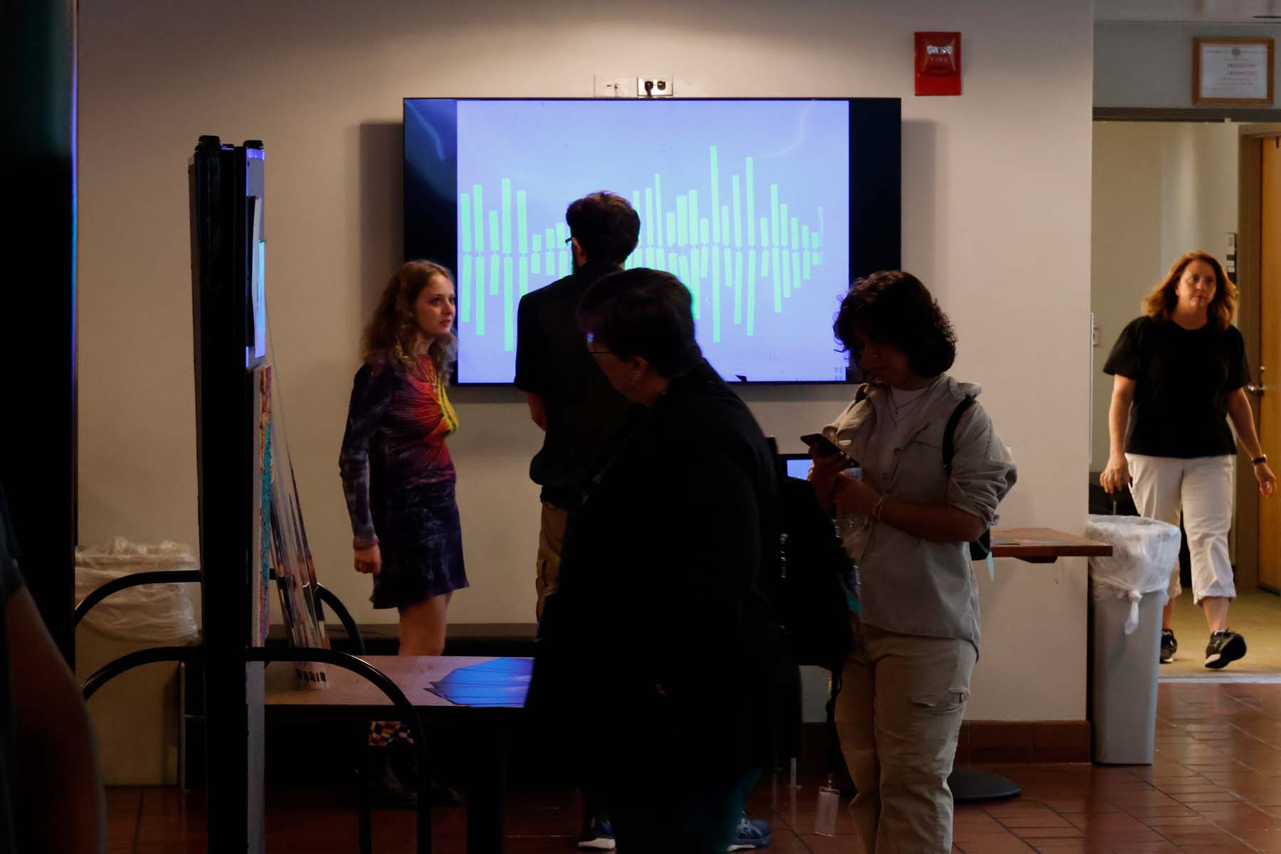 Three people examine a painting mounted to the while, while in the background a man looks inquistively at a screen displaying a waveform.