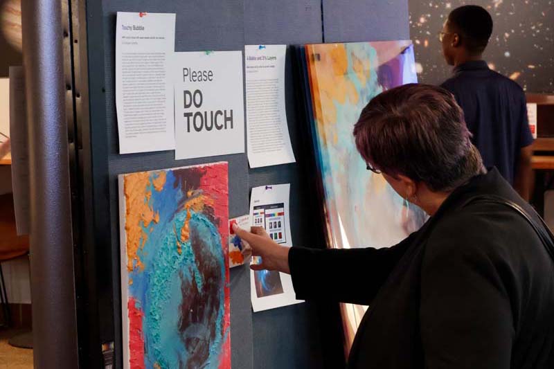 Two people examine a painting of a nebula with sign above it that says "PLEASE DO TOUCH". There are three paint swatches nearby that explain that each color (with corresponding texture) represent a different element present in the nebula.