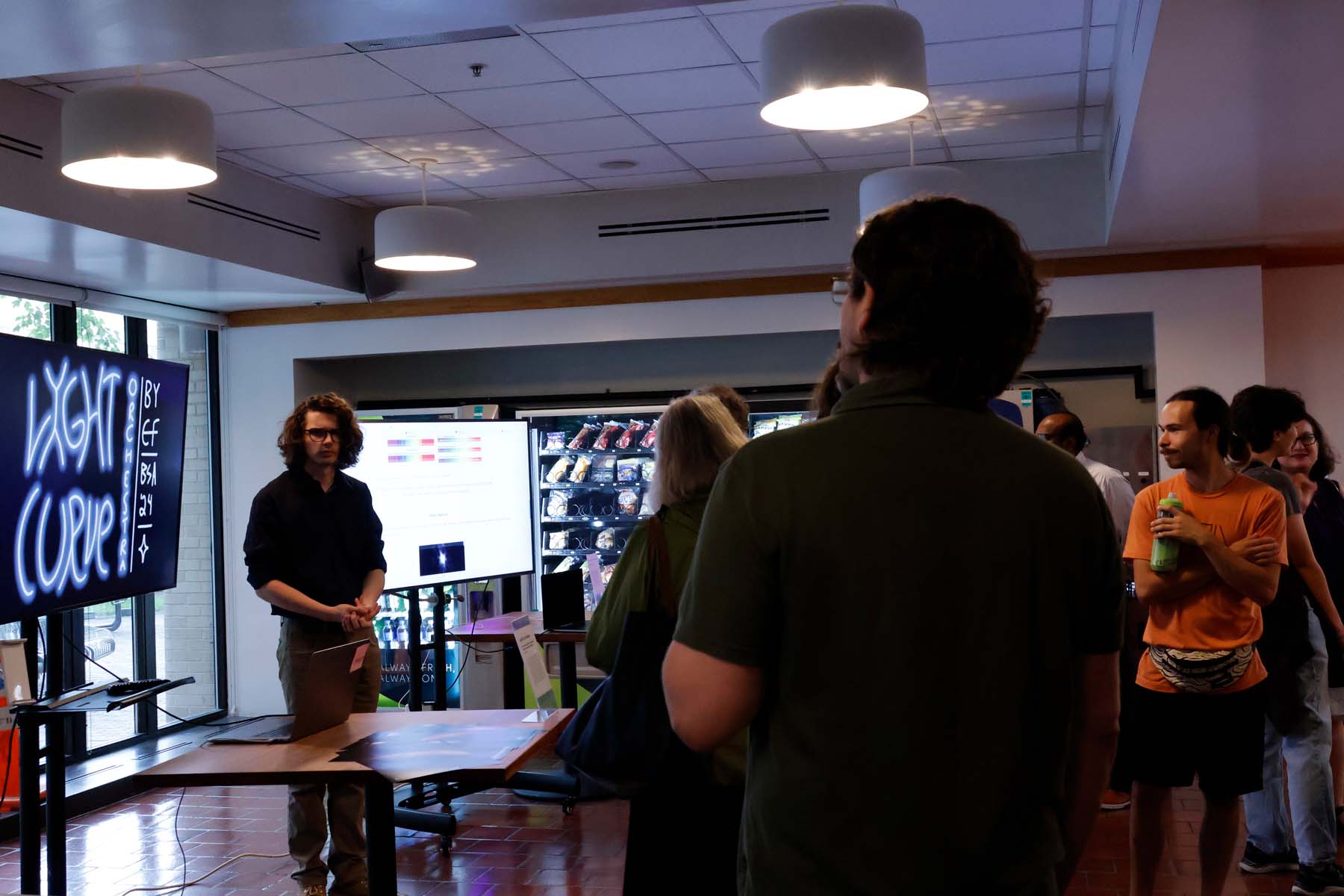 Two student projects are shown on large televisions: one is a website showing spectral data, the other an animation title screen reading "LIGHT CURVE ORCHESTRA".  A student artist stands between them, ready to take questions.