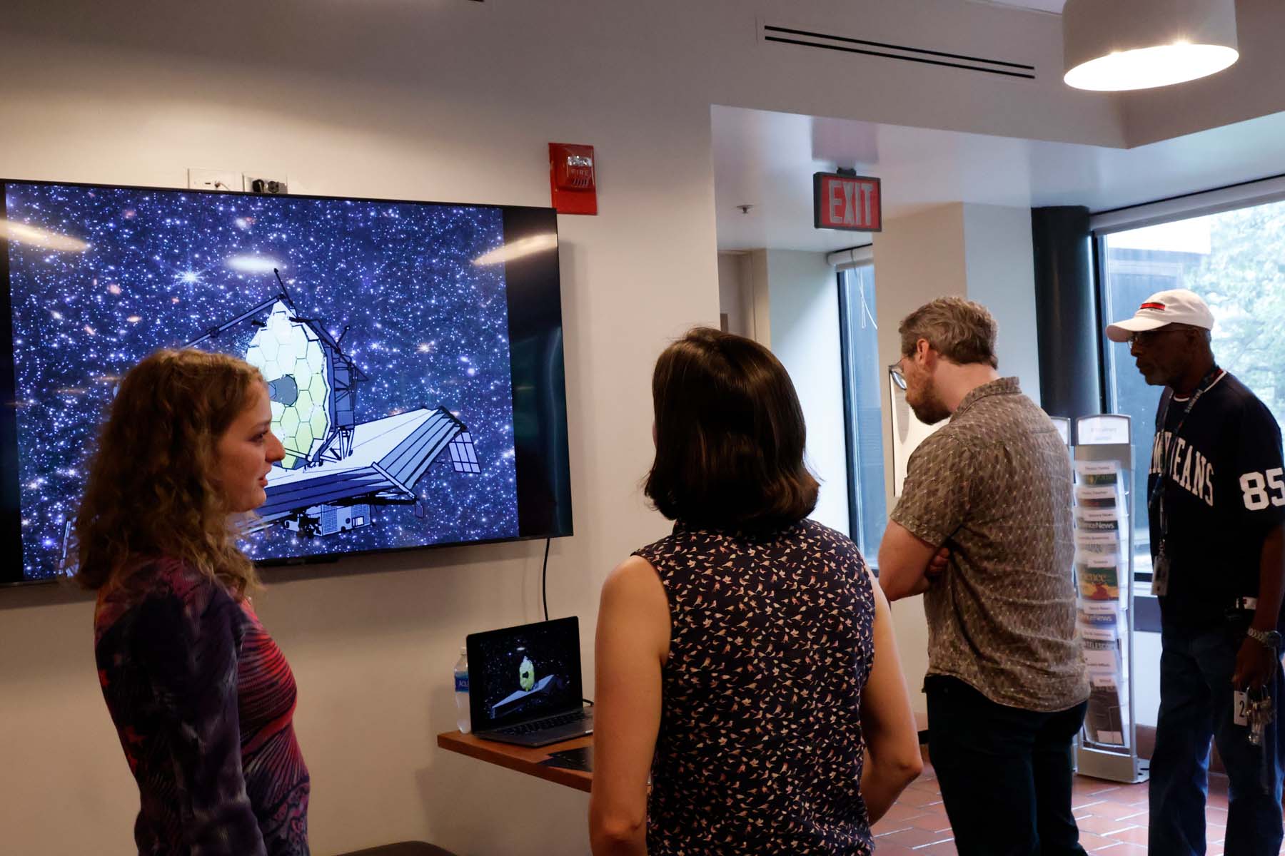 Photograph of a group watching an animation of JWST against a field of galaxies on a small laptop screen and mirrored on a large wall-mounted television. The artist stands to the left, discussing the project.
