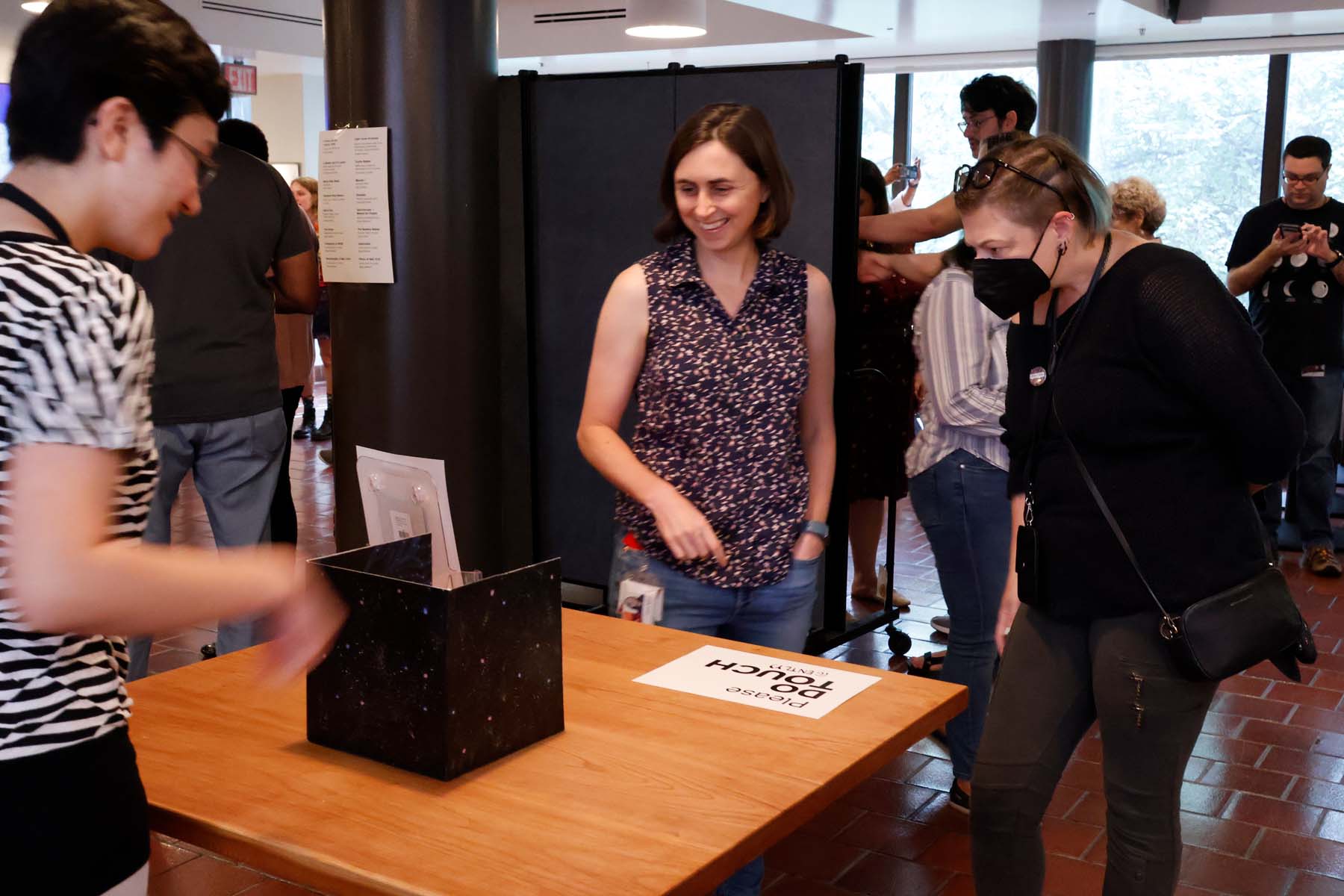 A black box sculptural piece sits on a table labeled “Please DO TOUCH (GENTLY)”. Two viewers are smiling, leaning in to look at the piece and speak with the artist, who is shown with her back to the camera.