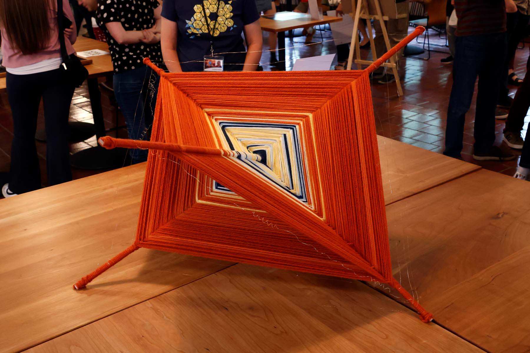 A yarn sculpture: Two wooden pieces form a cross and yellow and orange yarn is wrapped along back and forth forming a 3-dimensional diamond